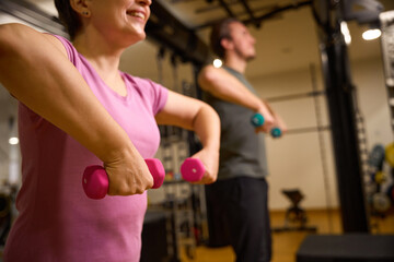 Mature woman and blurred trainer doing exercise with dumbbells for shoulders