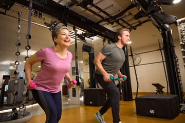 Trainer teaching mature smiling woman doing sport exercise with dumbbells in gym