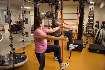 Trainer teaching mature woman doing exercise with sports sticks in gym