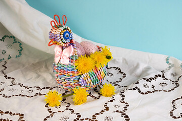 A rainbow chicken woven from a paper vine, with Easter eggs inside and yellow dandelions. He stands on a white lace napkin on a blue background. The concept of Easter