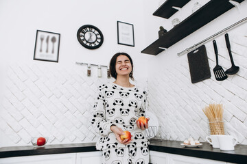 Cheerful woman standing in bright kitchen smiling laughing eating apple fruit, dressed in patterned...