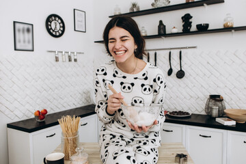 Cheerful woman sitting in bright kitchen mixing dough in a bowl, making homemade cookies, dressed in patterned pajama set. The kitchen counter arranged with ingredients, utensils, atmosphere domestic