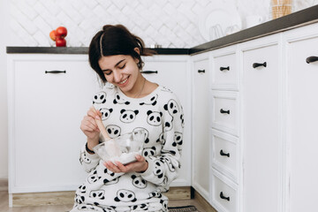 Cheerful woman sitting in bright kitchen mixing dough in a bowl, making homemade cookies, dressed in patterned pajama set. The kitchen counter arranged with ingredients, utensils, atmosphere domestic
