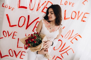 Woman sitting on chair with kisses lipstick is holding a bouquet of red roses, drawings of the word LOVE hand-painted, Valentine`s day concept celebration decoration. Love, holiday, party
