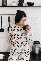 Cheerful woman standing in bright kitchen drinking hot beverage coffee tea, dressed in patterned...