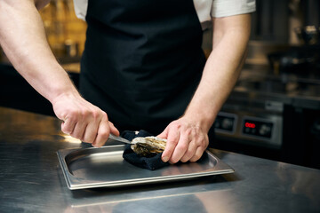 Cropped chef opening oyster with knife on iron tray for cooking dish at table