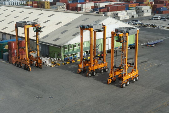 Bridgetown, Barbados 02 24 2024: Three orange straddle carriers in Bridgetown container terminal parking near warehouse and gantry cranes. There are many of other boxes from various shippers.