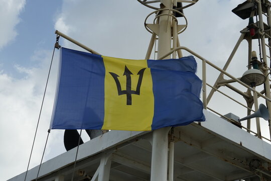 Flying Barbados flag, vertical triband of ultramarine and gold with the black trident-head, on the wind hanging on navigational mast near funnel of merchant container vessel when mooring in Bridgetown