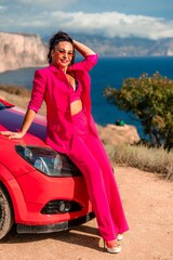 A woman in a pink suit is sitting on a red car. She is smiling and posing for the camera. The scene has a casual and relaxed vibe, with the woman enjoying her time outdoors.