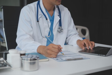 A professional and focused Asian female doctor in scrubs is working and reading medical research on her laptop in her office at a hospital.