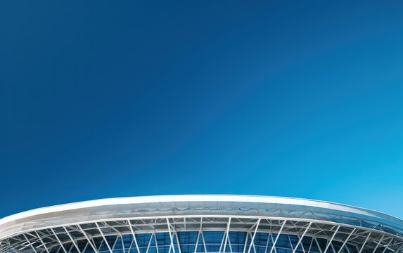 An architectural marvel, the image showcases the intricate design and structure of a modern stadium's roof against a cloudless blue sky