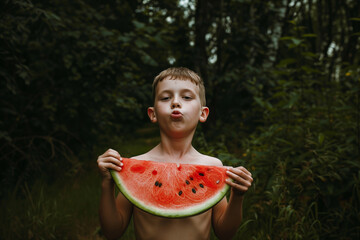 A young boy playfully puckers up while holding a large slice of watermelon, surrounded by the lush greenery of a forest