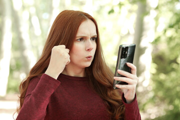 Woman with long red hair raises her fist angry because she sees something on her phone that makes...