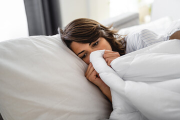 Young sick woman wrapped up in a blanket up to her nose lying in bed. Female person suffering from...