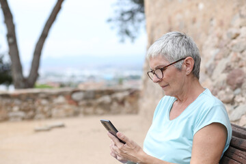 Obraz premium Mature woman with gray hair and glasses looks at her smartphone sitting in a park in summer