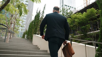 Back view of caucasian manager with formal suit walking up stairs while holding his suitcase in...