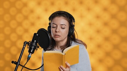 Narrator wearing headset reading aloud from book into mic against yellow background. Upbeat...