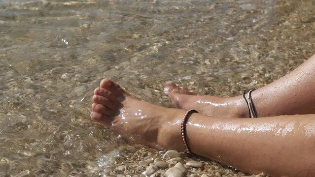CLOSE UP: Lady dips her bare feet, adorned with simple bracelets, in crystal clear shallow water at pebble shore on a sunny day. She is relaxing on the beach to the sound of gently lapping sea waves.