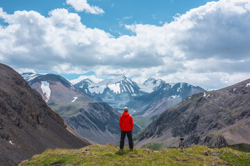 Man in vivid red jacket admire alpine scenery on sunlit green grassy hill near precipice edge. Guy on mountain pass enjoying view to few big snowy pointy peaks. Three large snow peaked tops far away.