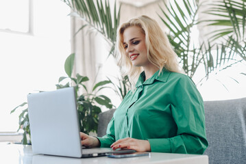 young focused freelancer working with notebook in cafe, typing text and smiling, concentrated caucasian blonde businesswoman in green shirt, green plants in background, freelance concept