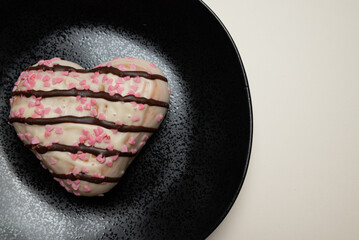 A heart-shaped donut with pink icing and sprinkles sits on a black plate. The plate is placed on a...