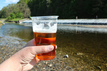 A cup with beer in hand in summer by the water, in the background a river with a weir, summer comfort