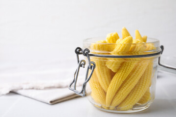 Tasty fresh yellow baby corns in glass jar on white tiled table, closeup. Space for text