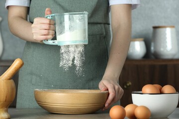 Woman sieving flour into bowl at table in kitchen, closeup