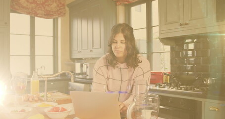 Image of flickering lights over woman eating in kitchen using laptop in kitchen at home