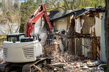 Fototapeta premium Demolishing an old home in a residential neighborhood to clear a lot and make way for a new home, heavy equipment with jawbone bucket to rip apart structure. 