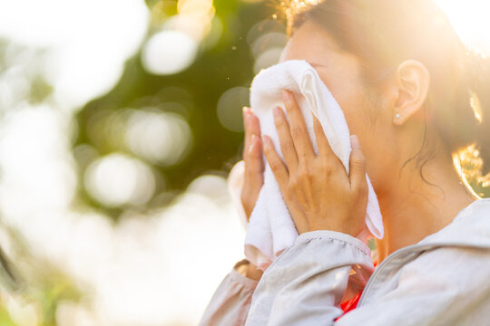Asian woman in sportswear wiping sweat on her face with towel during jogging exercise at public park at summer sunset. Healthy girl enjoy outdoor lifestyle sport training workout running in the city.