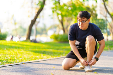 Asian man in sportswear tying running shoelaces during jogging exercise at public park in summer...