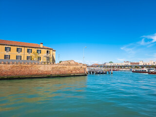 The majestic ancient building stands tall on the river, surrounded by boats and city buildings. Its reflection shimmers in the calm water under a clear blue sky, creating a serene and picturesque 