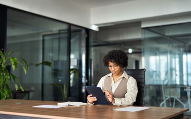 Young busy professional African American business woman company executive or entrepreneur using digital tablet working sitting at desk in office looking at tab, managing financial project data.