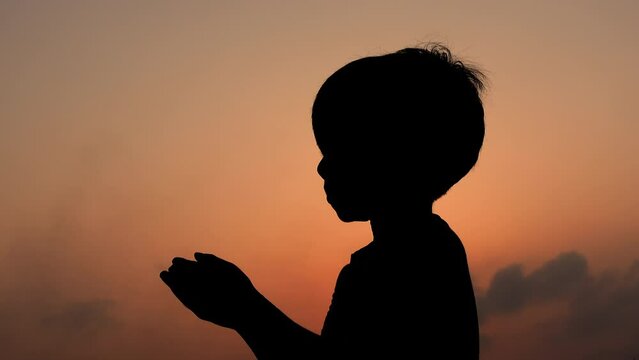A Muslim boy praying with his hands raised to the sky in the afternoon twilight. Silhouette Muslim boy carrying tasbeeh and praying on sunset time.