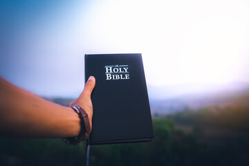 raised hand holding the holy Bible. background with blue sky in morning.