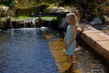 A young blonde girl in a blue dress stands in the shallows of a billabong