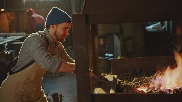 Professional blacksmith in apron and protective gloves heating iron rod on fire in charcoal forge when working in smithy