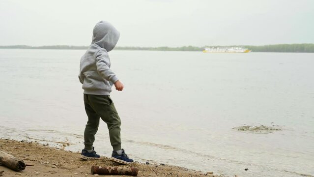 A small boy in a hoodie deftly throws a pebble into the water on the river bank. A child plays outdoors. Family active lifestyle. Connection with nature