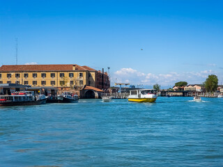 Boats floating in a canal in Venice. The waterway is surrounded by buildings, with a beautiful blue sky and clouds overhead. The boats are used for travel and are part of the vibrant harbor landscape