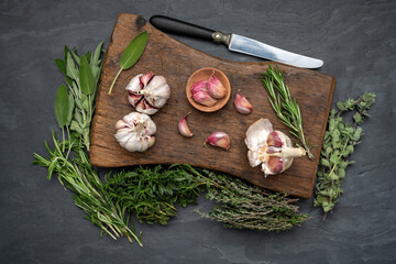 Still life, garlic, fresh green herbs and a knife on a rustic wooden cutting board on a black slate...