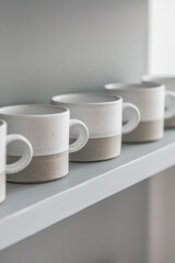 A series of minimalist ceramic or porcelain mugs with clean lines and matte finishes, arranged in a row on a solid-colored shelf, inviting relaxation and indulgence with every sip. 