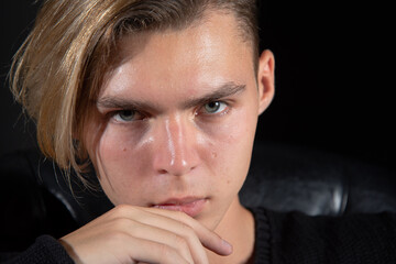A young attractive guy is sad while sitting on a chair. Black background.