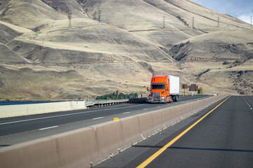 Bright orange bonnet big rig semi-truck with grille guard transporting cargo in dry van semi-trailer climbing uphill on the highway road with bare bald mountains