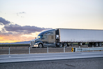 Dark gray big rig semi-truck tractor transporting cargo in refrigerator semi trailer driving on the divided highway road with sunset sky in California