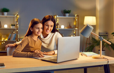 Young cheerful happy mother with her teen school girl daughter studying online class on laptop at...