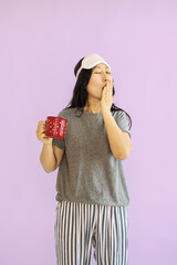 Young Asian woman yawning while holding a cup of tea or coffee on purple background