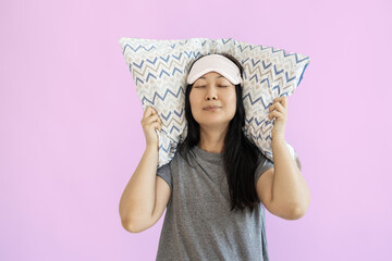 happy young Asian woman wears pyjamas jam sleep eye mask rest at home hold pillow isolated on pink