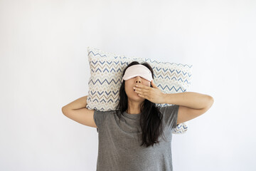 Young tired Asian woman with sleeping mask and pillow yawning on white background