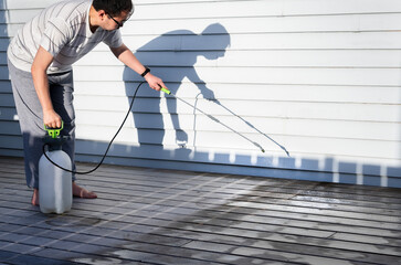 Man cleaning wooden deck with hand-held pressure sprayer, shadow on the wall in the afternoon sun.